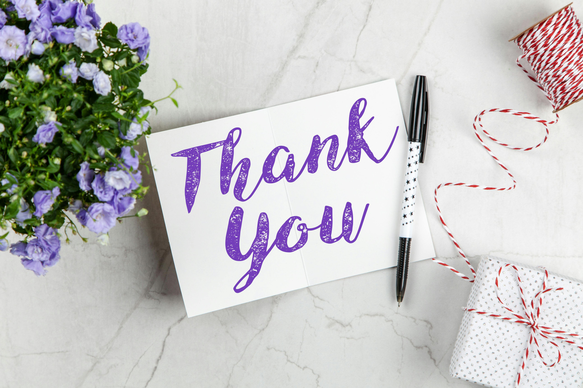 A white card is on a table top with the words thank you written in purple. To the left of the card is a poses of purple flowers and to the right a small wrapped gift with red string tied around.