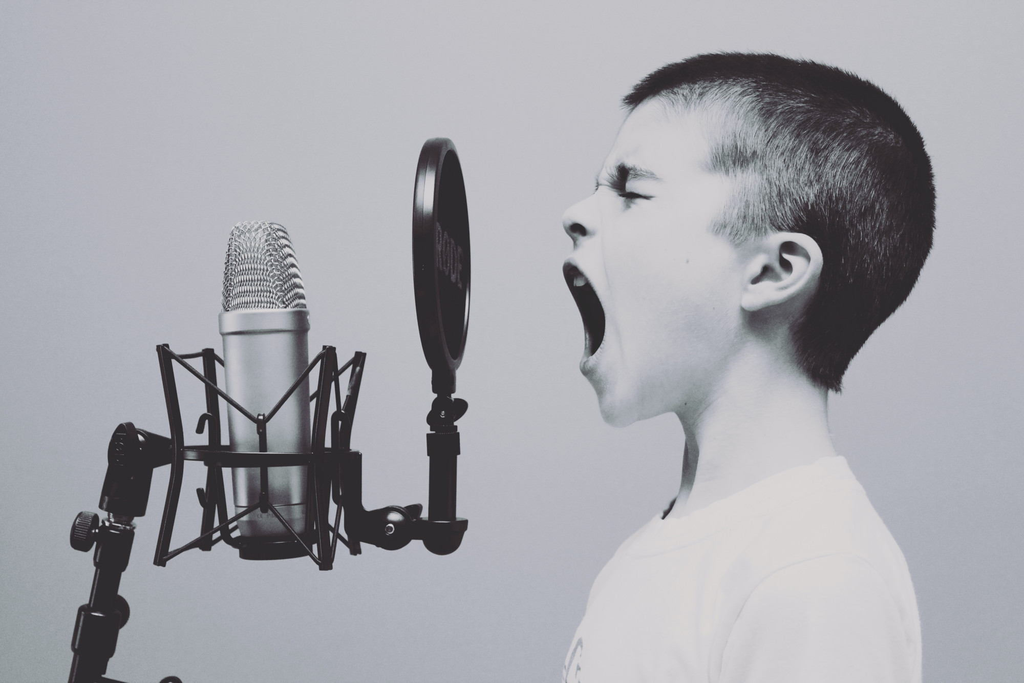 Black and white image of young boy stood in front of a microphone with his mouth wide open as if to scream to be heard.