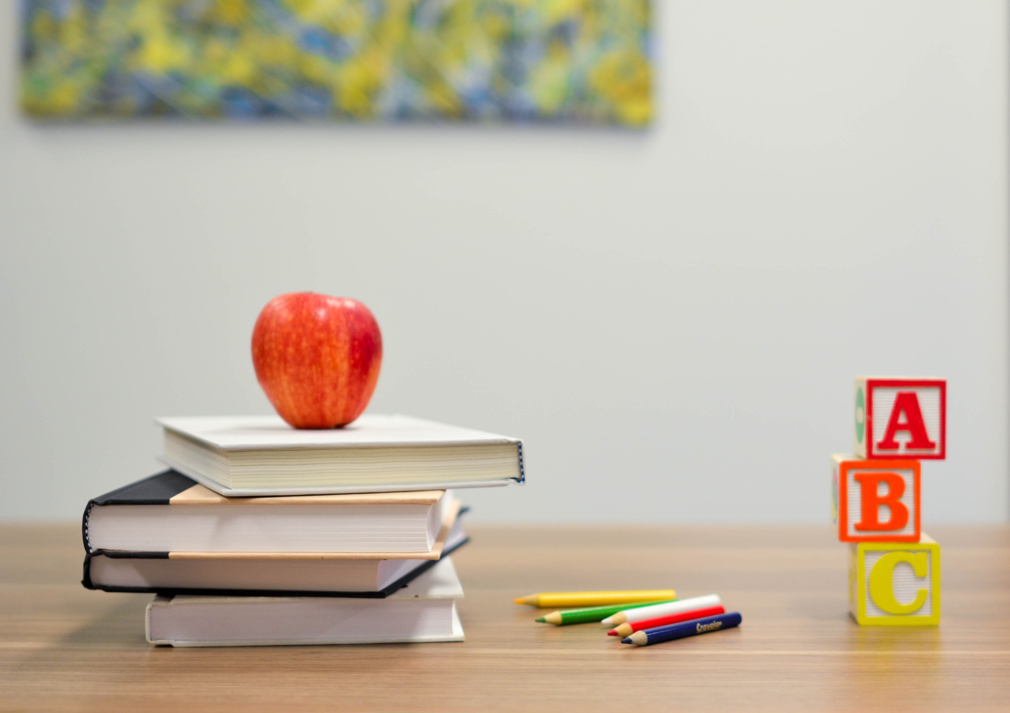 A table top is bottom of the picture upon sits a stack of 4 books with a red apple on top of them. To the right of them is some coloured pencils and to the right of those is three building blocks stacked with the letters ABC