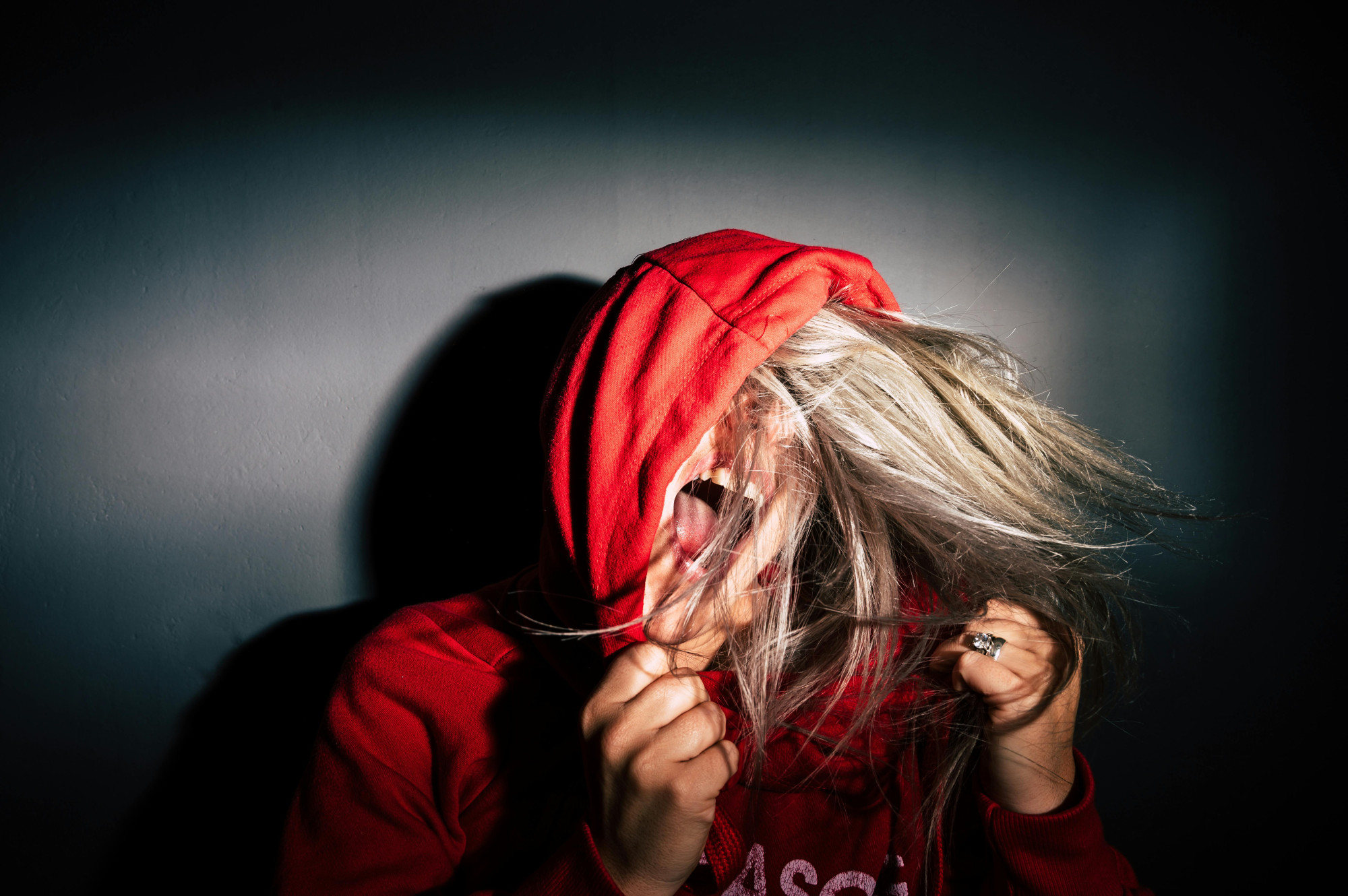 Teen girl in red hoodie and darkened room with her hood up and face hidden by her hair seemingly screaming into the empty room