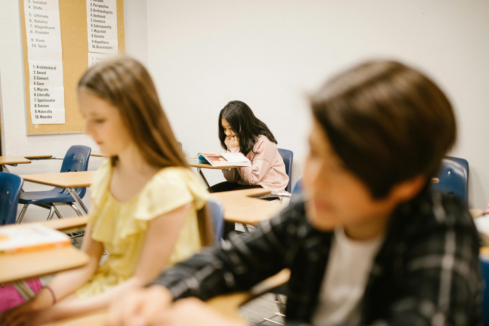 Image of three children in a classroom at separate desks. The two children to the front are blurred in the image but doing school work. The girl sat at the desk in the of the room is hunched over her school book looking worried with her hands scrunched near her mouth 